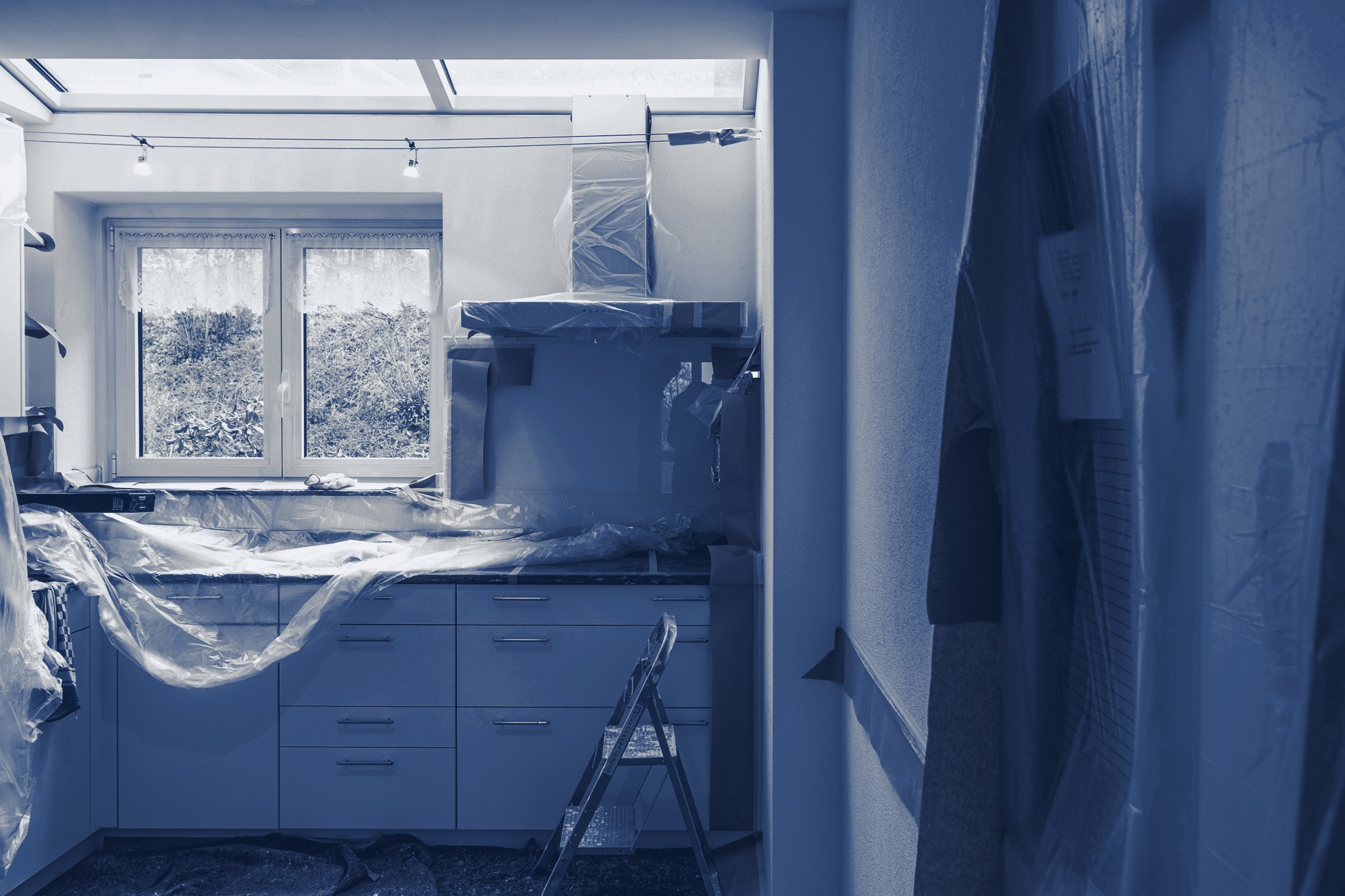 Apartment kitchen under renovation: new white cabinets and hood covered in plastic and paper, step ladder, tinted navy