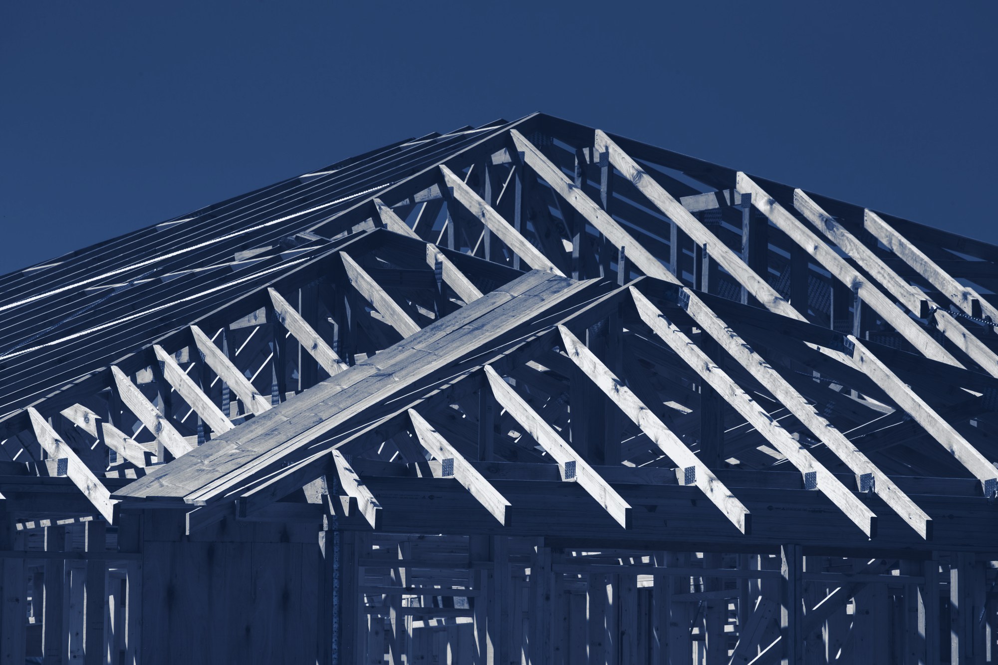 New house under construction: timber wall framing and roof trusses against a blue sky, tinted navy