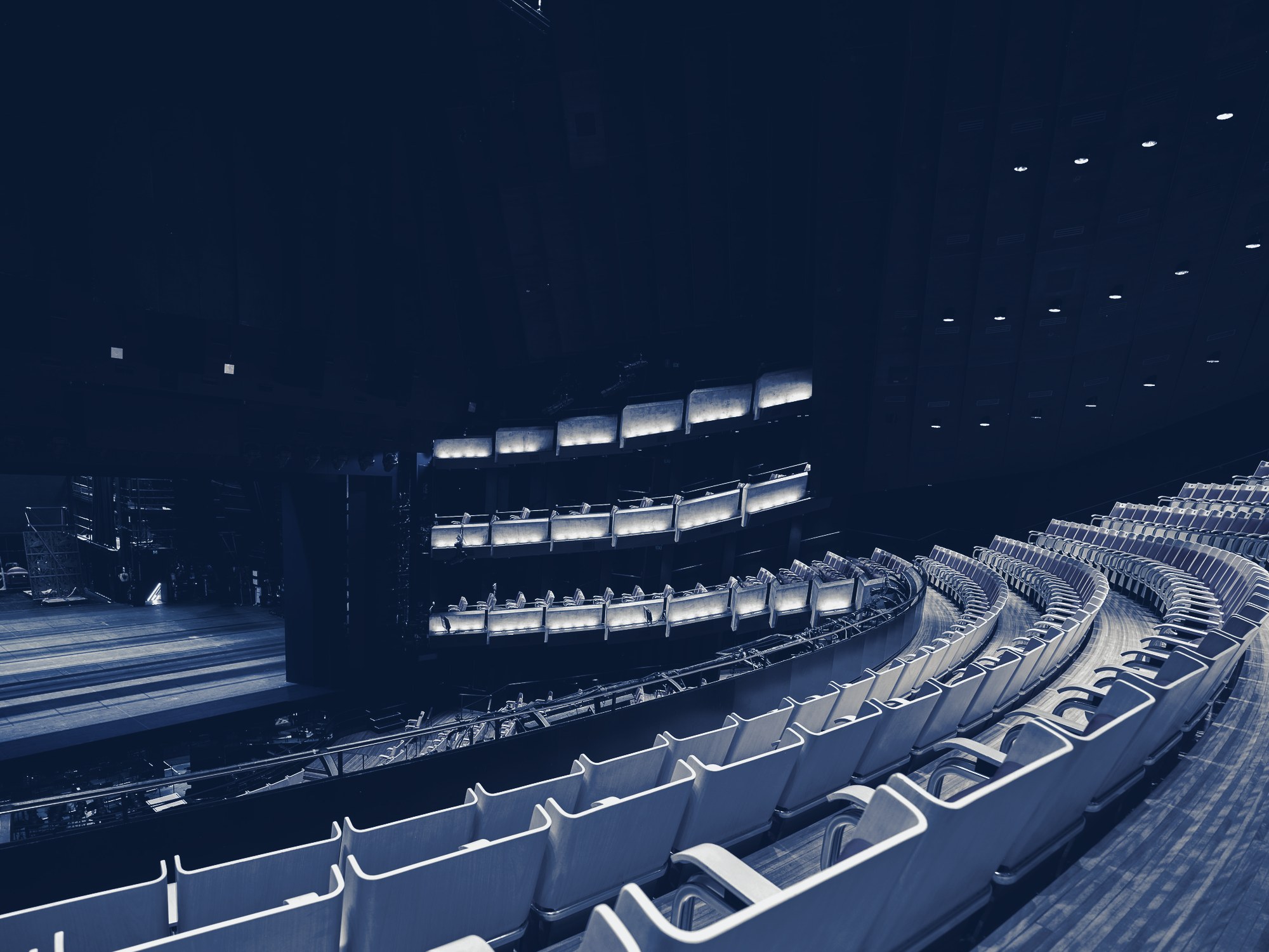 Empty concert hall/theater interior with curved rows of seats, balconies and a lit stage, tinted navy