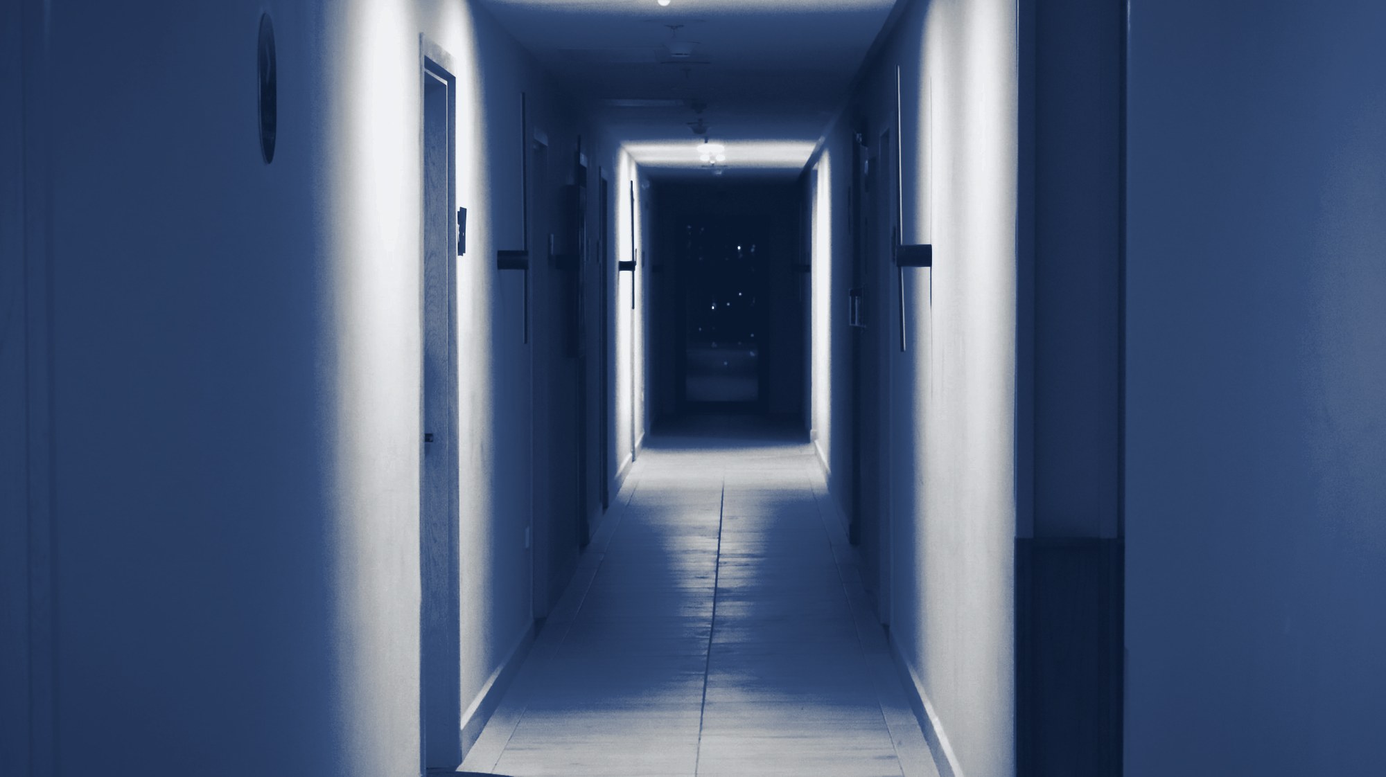 Long, dimly lit hotel-style corridor with room doors on both sides, tinted navy