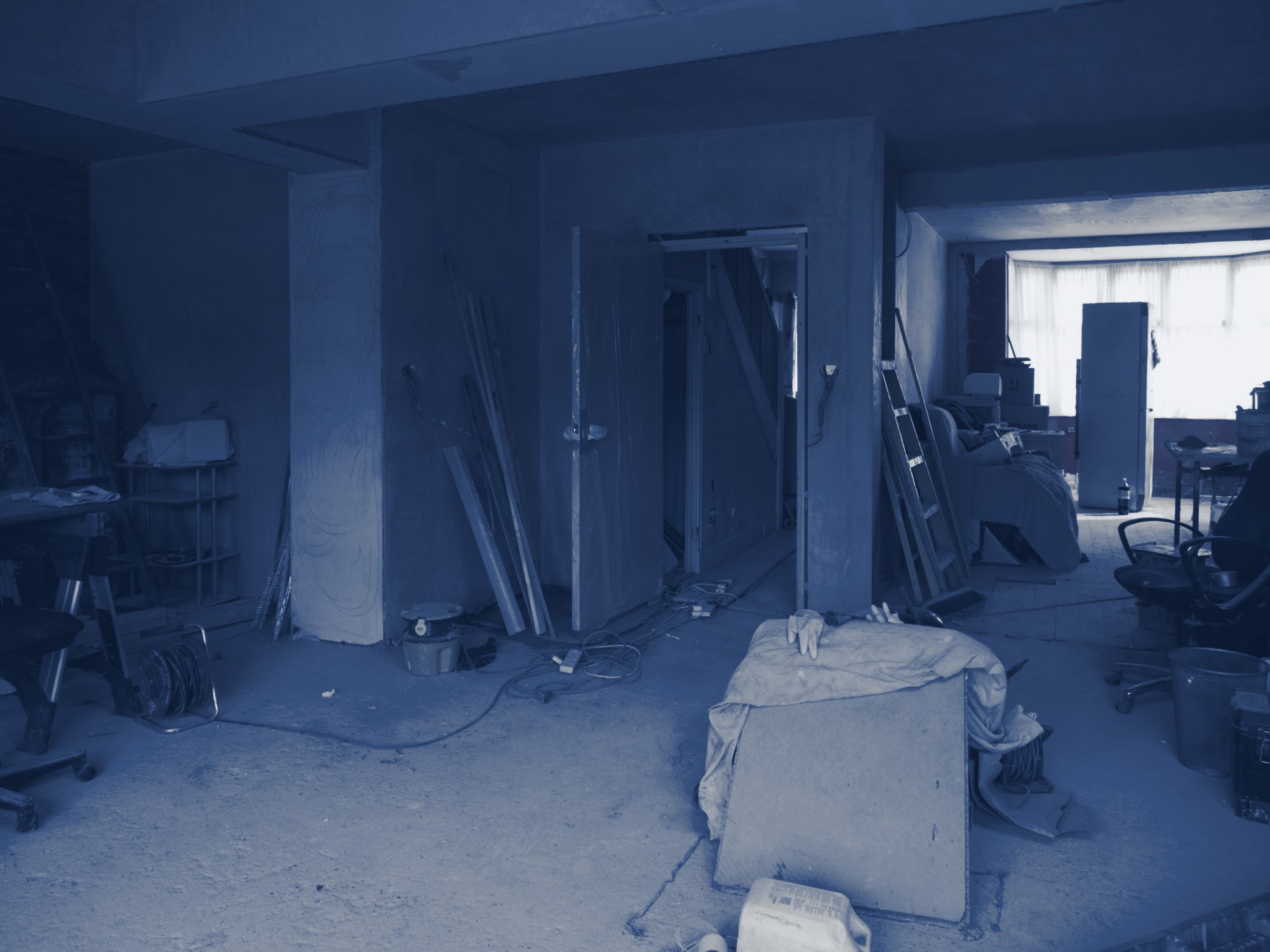 Building interior stripped for renovation: bare plaster walls, door frames, ladders, cable reels and dust sheets, tinted navy