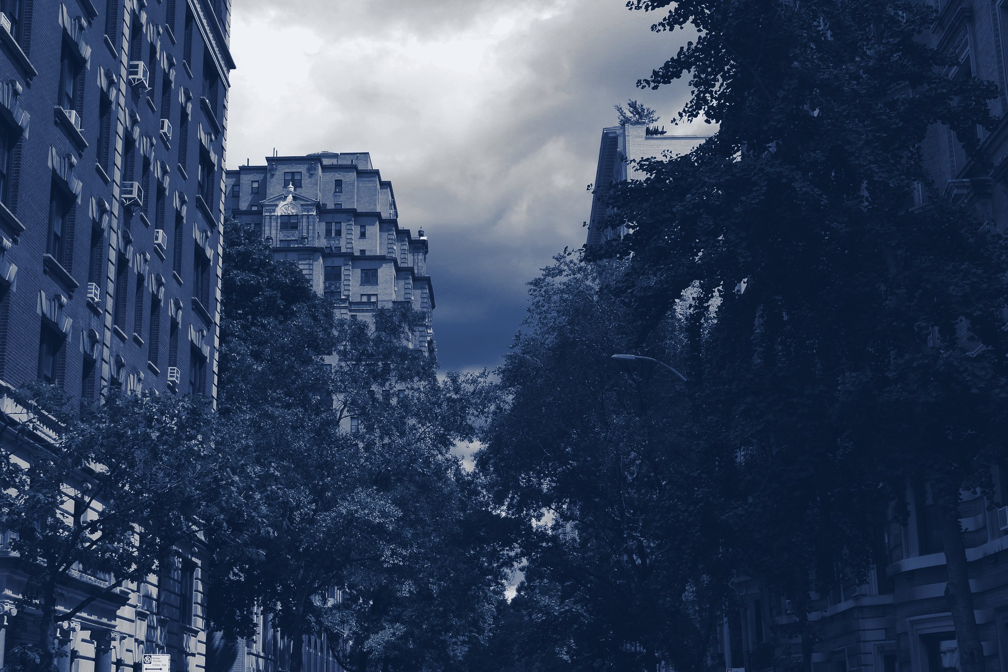 Pre-war mid-rise apartment buildings along a street lined with mature trees, cloudy sky, tinted navy